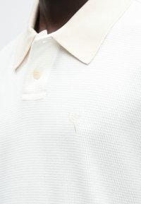 Detailní záběr osoby s bílým texturovaným polokošilím s béžovým límcem a jemným srdcovým emblémem na hrudi.