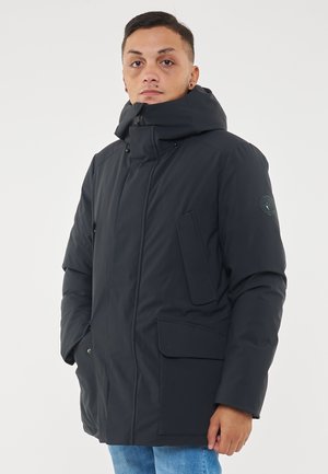 Parka isolato di colore blu scuro con colletto alto e cappuccio regolabile. Presenta molteplici tasche frontali e un logo circolare sulla manica sinistra.