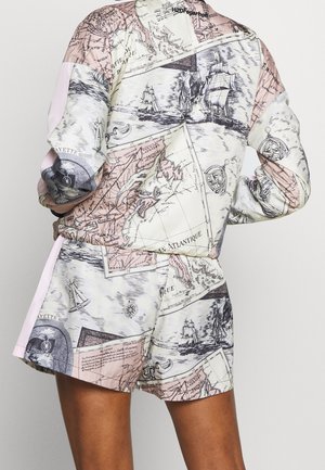 Osoba w dopasowanych szortach i bluzce z długim rękawem z nadrukiem starej mapy i żaglowca w miękkich różowych i kremowych odcieniach.