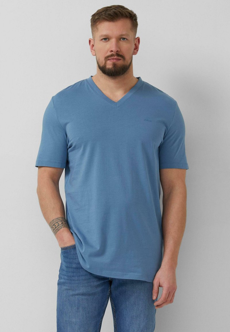 Homme avec barbe et tatouage portant un t-shirt bleu à col en V et manches courtes, et un jean bleu, debout avec une main dans la poche.