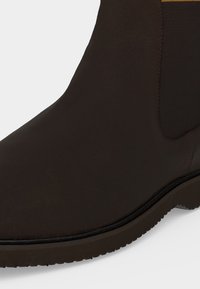 Gros plan d'une botte Chelsea marron foncé montrant une tige en cuir lisse, un panneau latéral élastique et un bord de semelle rainuré.