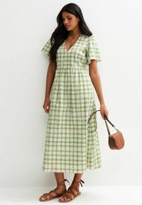 Grün-weiß karierter Maxikleid mit V-Ausschnitt, kurzen Puffärmeln und elastischem Bund; kombiniert mit einer braunen Umhängetasche und Sandalen.