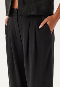 Pantalons plissés noirs en tissu texturé, avec une fermeture à bouton et des poches latérales, montrés de la taille jusqu'en bas.