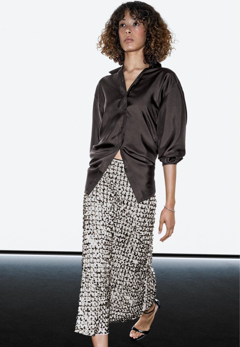 Vrouw met krullend haar loopt, draagt een glanzend donker overhemd, een wit-zwart patroonrok en zwarte sandalen met open tenen.