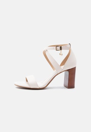 Sandale à talon haut en cuir blanc avec talon bloc en bois, bride à la cheville et charm en forme de boucle dorée sur fond gris clair.