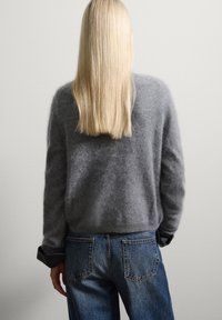 Grauer, flauschiger Pullover mit langen Ärmeln, kombiniert mit dunkelblauen hochgeschnittenen Jeans, von hinten vor einem hellgrauen Hintergrund betrachtet.