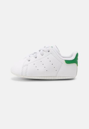 Biały dziecięcy sneaker z białymi sznurówkami, perforowanymi bocznymi detalami oraz zielonym paskiem na pięcie z logo na jasnym tle.