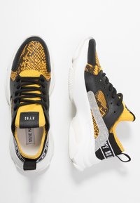 Baskets avec une tige à motif serpent noir et jaune, semelle extérieure blanche et accents argentés. Comprend des lacets noirs et un col rembourré.