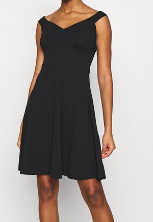 Vrouw draagt een mouwloze zwarte jurk met een aansluitend lijfje en een uitlopende rok, tegen een effen witte achtergrond.