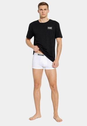 Uomo che indossa una maglietta nera Moschino sollevando l'orlo per mostrare i boxer bianchi Moschino, in piedi scalzo su sfondo semplice.