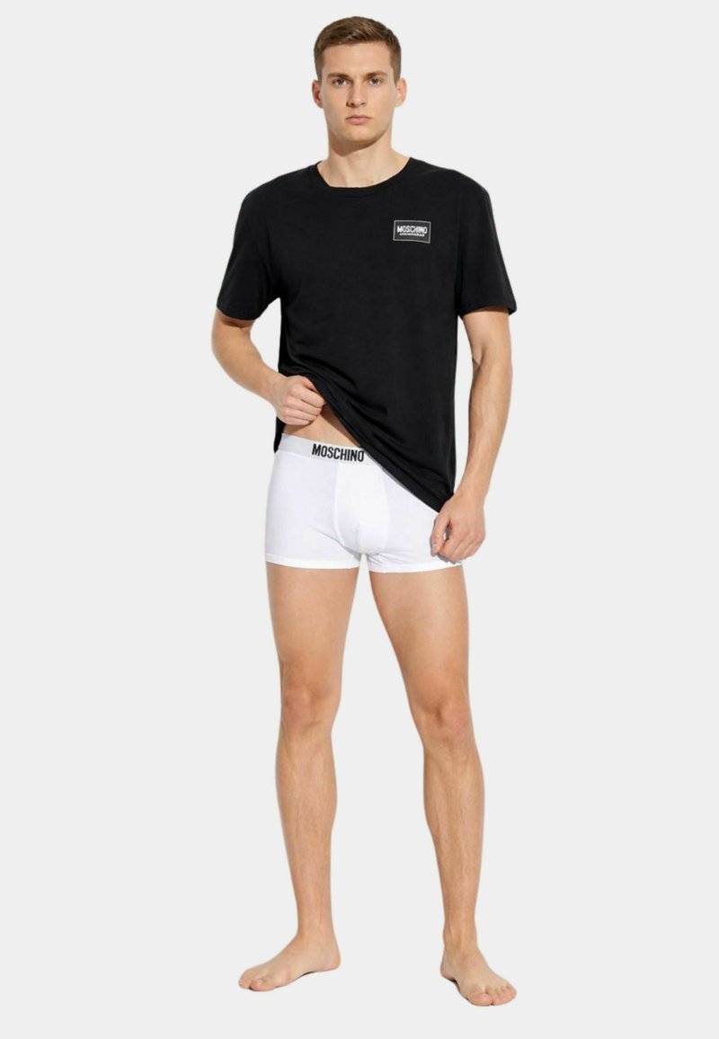 Uomo che indossa una maglietta nera Moschino sollevando l'orlo per mostrare i boxer bianchi Moschino, in piedi scalzo su sfondo semplice.