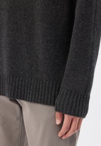 Dunkelgrauer Pullover mit weicher Textur, gerippten Bündchen und Saum. Über hellgrauen Hosen getragen, zeigt eine Nahaufnahme des Ärmels und der Hand.