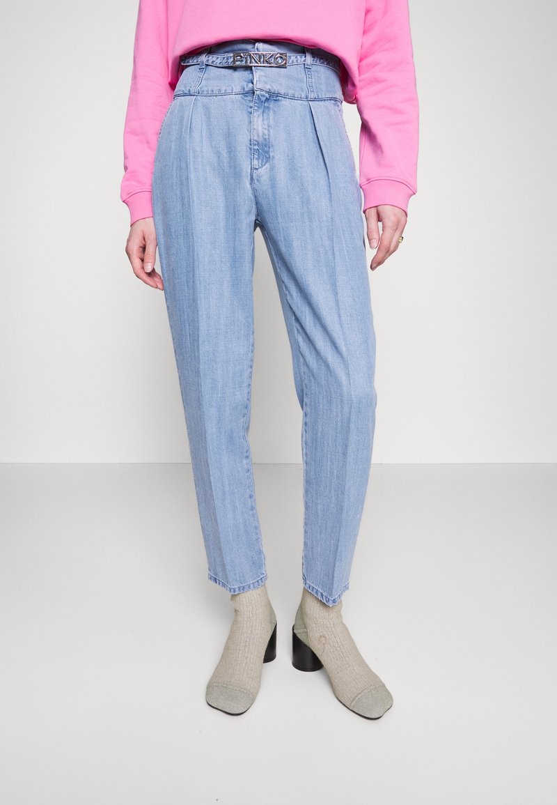 Pantalons en denim plissés de taille haute, bleu clair, avec une ceinture assortie, associés à des chaussettes grises jusqu'au genou et des chaussures à talons noires.