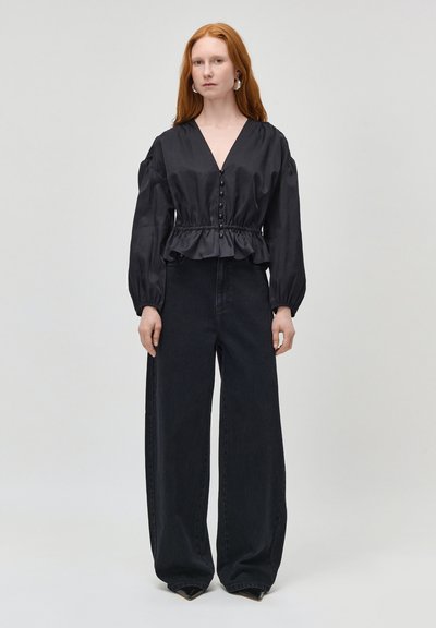 Femme aux longs cheveux roux portant un chemisier noir à manches longues boutonné avec une taille à volants et un jean noir à jambes larges, debout devant un fond uni.