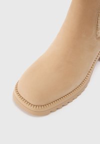 Botine din suede bej, cu vârf rotunjit și un panou lateral texturat. Sulița este maro cu detalii în model zigzag.
