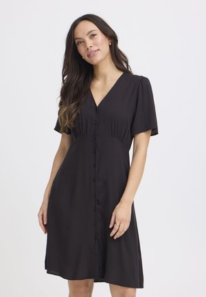 Kobieta o długich ciemnych włosach, ubrana w czarną sukienkę na guziki z krótkimi rękawami, stojąca na tle jednolitego białego tła.