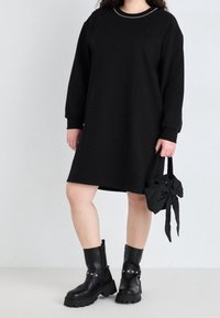 Robe pull oversized noire à manches longues, avec un col rond orné de perles. Associée à des bottes noires à lacets et un petit sac agrémenté d'un nœud.