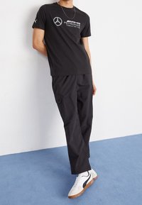 Czarny t-shirt z krótkim rękawem z białym logo Mercedesa i napisem, połączony z czarnymi spodniami cargo i białymi trampkami z brązowymi podeszwami.