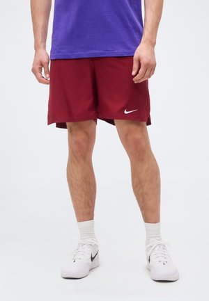 Mężczyzna ubrany w czerwone krótkie spodenki Nike, białe sneakersy Nike, białe skarpetki i fioletową koszulkę, stojący na tle prostego jasnego tła.