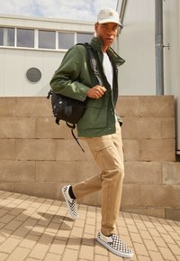 Grønn parkajakke, hvit T-skjorte, khaki cargo bukser og sort-hvite rutete slip-on sko. Svart ryggsekk, utendørs miljø.