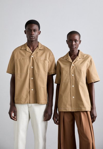 Krátké rukávy, tan košile s předními kapsami a knoflíkovými zapínáními na modelu; spárované se světlými a hnědými šitými kalhotami. Hladká, jednolitá tkanina.