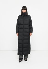 Dlhý čierny puffer kabát s vysokým golierom, obsahujúci plný predný zips a horizontálne prešívané časti. Nosený cez tmavé džínsy a čierne topánky.