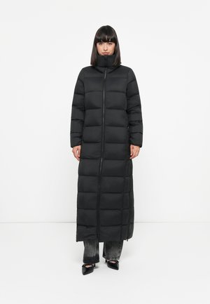 Lange zwarte pufferjas met hoge kraag, voorzien van een volledige voorkantrits en horizontaal gestikte secties. Gedragen over donkere jeans en zwarte schoenen.