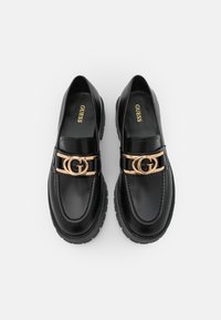 Mocassins en cuir noir avec un bout arrondi, un emblème double G doré, des détails cousus et une semelle en caoutchouc texturée. L'intérieur comporte le nom de la marque.