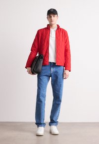 Rode gewatteerde jas met een hoge kraag, witte poloshirt, blauwe spijkerbroek en witte sneakers. Het model houdt een zwarte tas vast.