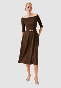 Abito marrone con scollo a barca, maniche a tre quarti, corpetto aderente e gonna svasata, caratterizzato da una cintura con accento metallico e tasche laterali.