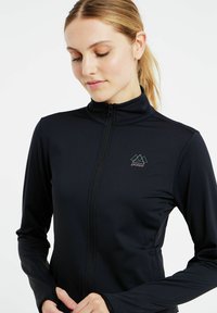 Schwarze Zip-Jacke aus dehnbarem Material. Sie verfügt über einen hohen Kragen, lange Ärmel und ein dezentes Logo mit geometrischen Akzenten auf der Brust.