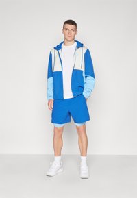 Chaqueta ligera azul con detalles grises, camiseta blanca y pantalones cortos azules. Tejido atlético con cordones y diseños de mangas en contraste.