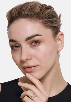 Close-up of a model with light skin, brown hair, wearing a black top. She has blue eyes and three silver rings on her fingers.