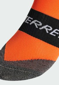 Calcetín naranja hasta el tobillo con franja horizontal negra y texto blanco. Cuenta con un patrón de punto texturizado y una zona del dedo del pie reforzada en gris.