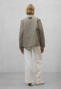 Geplüschte olivgrüne Jacke mit einem gerippten Kragen, oversized Schnitt und dezentem Branding auf der Rückseite, kombiniert mit weißen, weit geschnittenen Hosen.