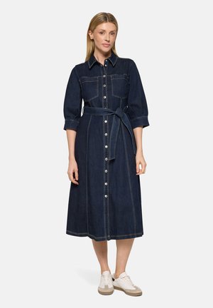 Kvinde iført en mørkeblå denim-midi-kjole med knapper foran, brystlommer, taljebælte, trekvartærmer og hvide sneakers på en ensfarvet baggrund.