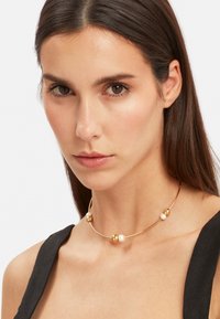 Guld chokersmykke med tre sfæriske accenter og to hvide perler. Glat metalfinish, minimalistisk design og fleksibel form.