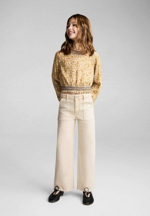 Haut à manches longues, à motif floral jaune avec taille froncée. Jean crème taille haute et large. Chaussures noires avec nœuds à l'avant.