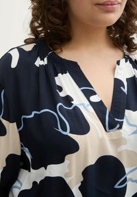 Leichte Bluse mit einem dunklen marineblauen und cremefarbenen abstrakten Muster, V-Ausschnitt und Raffung an den Schultern. Satinähnliche Textur.