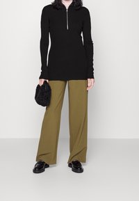 Svart zip-up långärmad top, olivgrön vida byxor, svart texturerad handväska och glänsande svarta loafers, på en ljusgrå bakgrund.