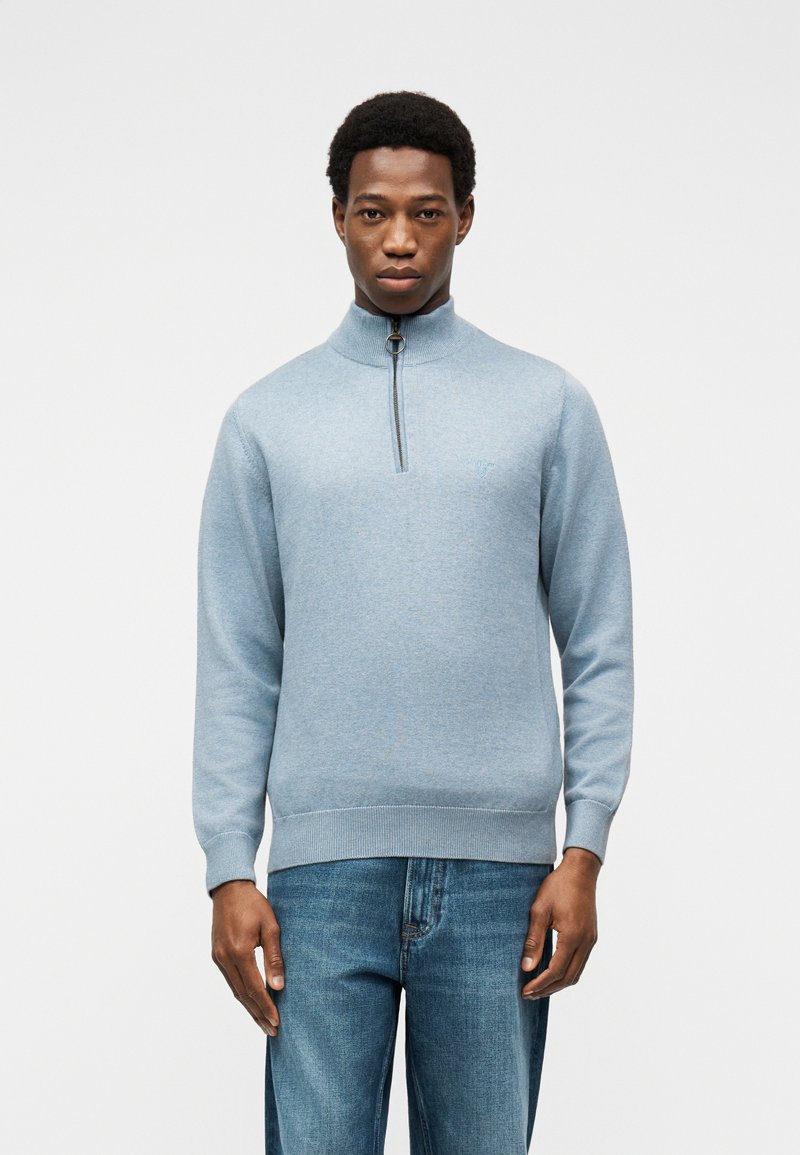 Jeune homme portant un pull bleu clair à fermeture éclair quart et un jean bleu, debout devant un fond blanc uni.