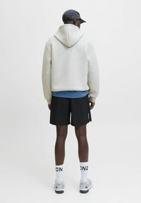 Person in einem hellgrauen Kapuzenpullover, schwarzen Shorts, weißen Socken mit "CNZ"-Text, grauen Sneakers und einer schwarzen Kappe, die mit dem Rücken zu einer einfarbigen Hintergrund steht.