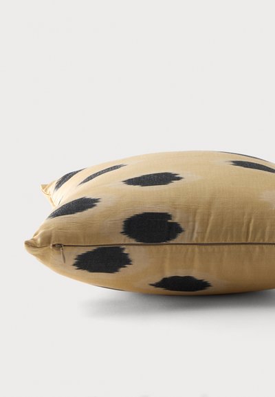 Coussin rectangulaire beige avec des taches noires irrégulières et une fermeture éclair visible sur le côté, reposant sur une surface claire.