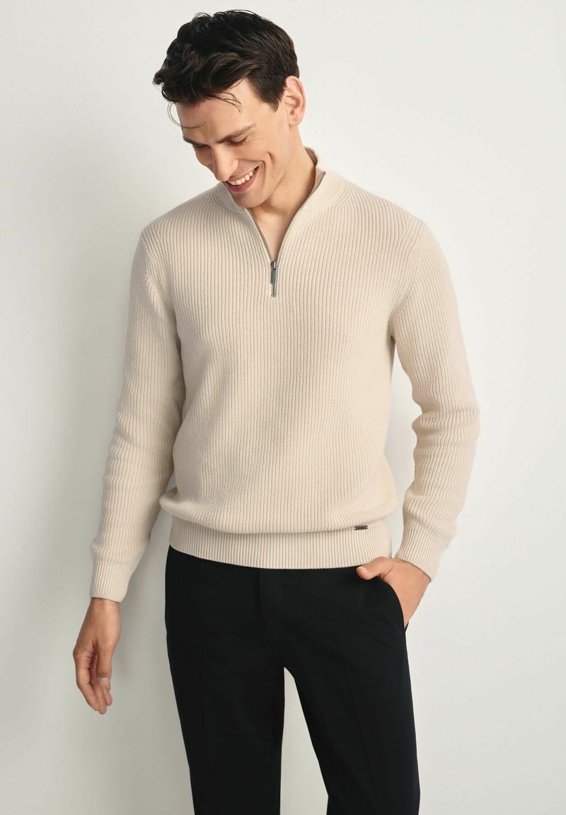 Beżowy prążkowany sweter z kołnierzem na pół-zamek i teksturowanym materiałem. Dopasowane mankiety i ściągacz w pasie. Połączony z czarnymi spodniami.