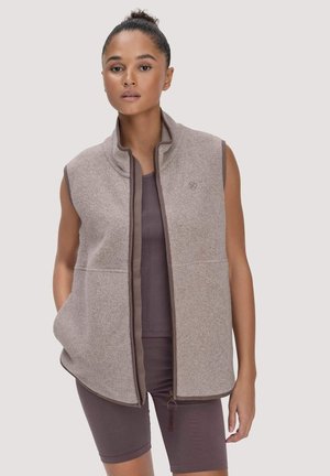 Frau trägt eine ärmellose Weste in Taupe mit Reißverschluss über passenden eng anliegenden Shorts und Top, steht mit einer Hand in der Westentasche.