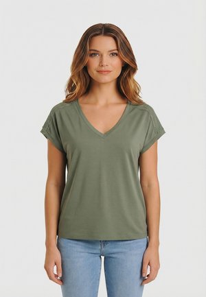 Joven con cabello castaño ondulado, vestida con una camiseta de cuello en V color verde oliva con detalles de encaje en los hombros y jeans azul claro, posando contra un fondo blanco.