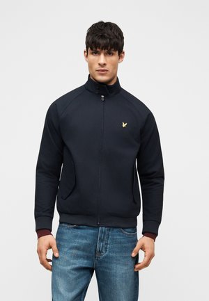 Άνδρας που φοράει μαύρο μπουφάν με φερμουάρ και χρυσό λογότυπο αετού στο στήθος και μπλε τζιν, στέκεται μπροστά σε απλό φωτεινό φόντο.