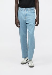 Hombre vistiendo jeans de pierna recta de color azul claro con cierre de botones y zapatillas blancas con franjas negras sobre un fondo blanco.