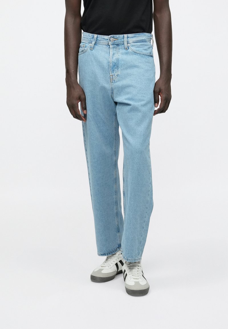 Hombre vistiendo jeans de pierna recta de color azul claro con cierre de botones y zapatillas blancas con franjas negras sobre un fondo blanco.