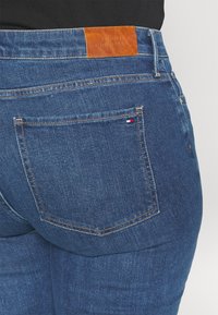 Gros plan sur la poche arrière d'un jean en denim bleu avec un petit logo Tommy Hilfiger et une étiquette de marque en cuir marron sur la taille.